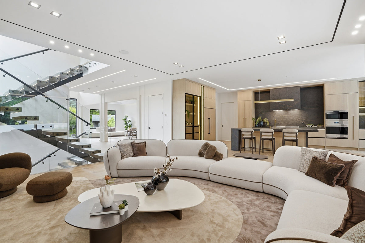 Modern living room with curved sectional sofa and round coffee tables, open to a kitchen and floating staircase.