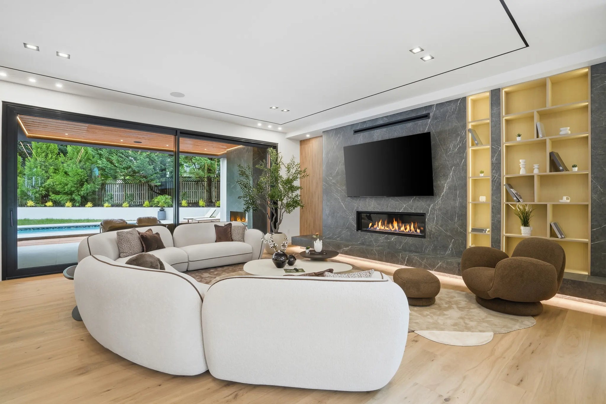 A modern living room with white curved sofas, a wooden coffee table, a fireplace, large windows overlooking a pool, and built-in wooden shelving.