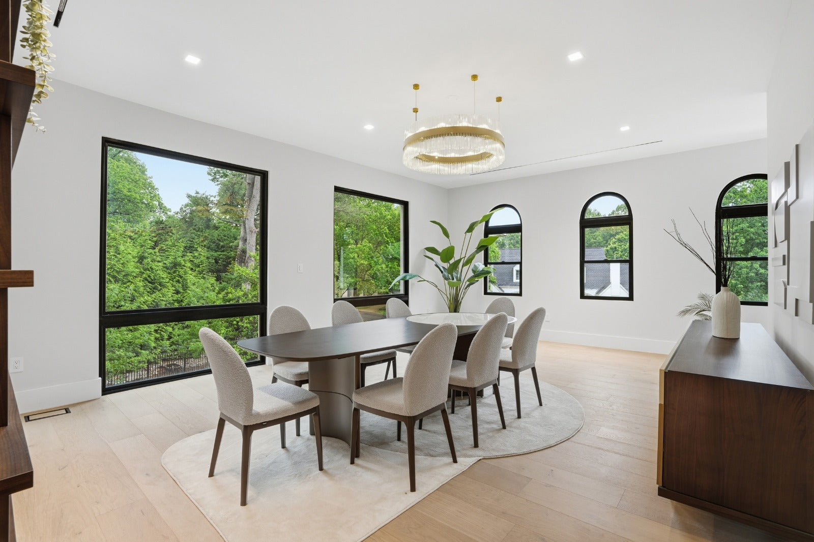 Bright dining room with a large wooden table, beige chairs, a circular chandelier, and floor-to-ceiling windows overlooking greenery.