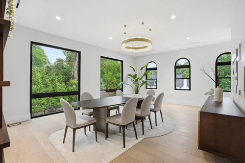 Bright dining room with a large wooden table, beige chairs, a circular chandelier, and floor-to-ceiling windows overlooking greenery.