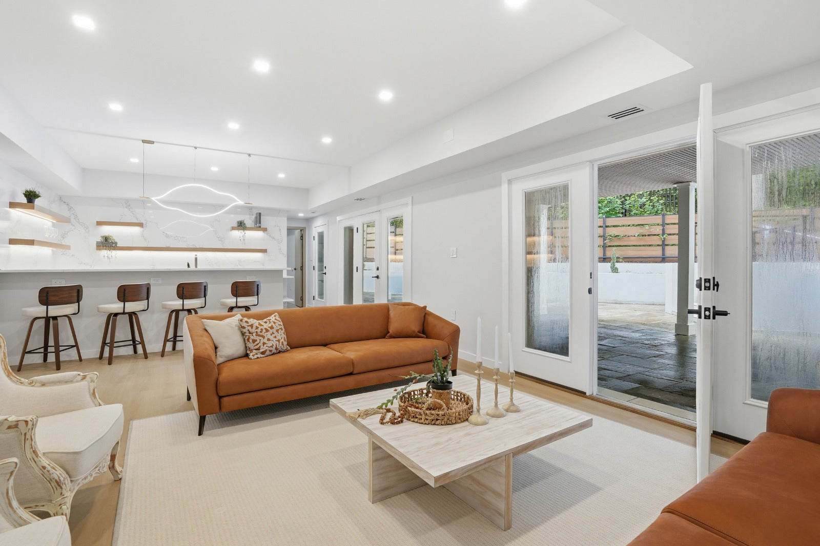 Modern living room with an orange sofa, white coffee table, and a kitchen area featuring bar stools and pendant lights, with glass doors leading to a patio and water view.