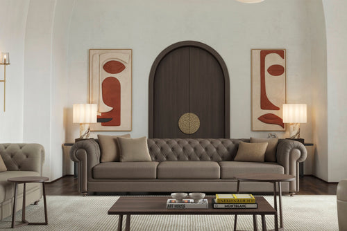 Elegant living room with a tufted brown Chesterfield sofa, wooden coffee table with books and decor, two armchairs, table lamps, and abstract art above a dark arched doorway.