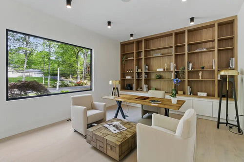 A modern living room with white armchairs, a wooden desk, shelves, and a large window overlooking greenery