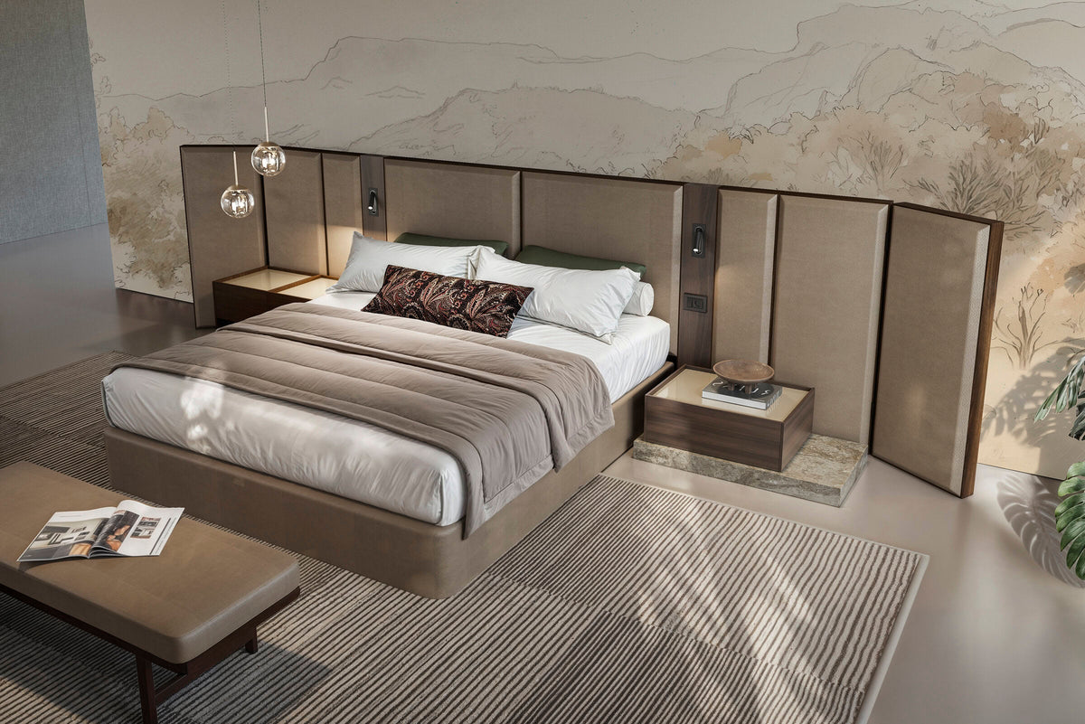 Modern bedroom with tall upholstered headboard, matching nightstands, low platform bed, and a bench at the foot of the bed against a textured stone-effect wall.